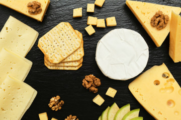 Pieces of cheese on a dark background. Cheeseboard. Sliced ​​apple and nuts on the board. Hard cheese, camembert, roquefort, emmental.