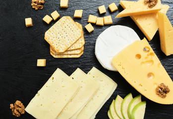 Pieces of cheese on a dark background. Cheeseboard. Sliced ​​apple and nuts on the board. Hard cheese, camembert, roquefort, emmental.