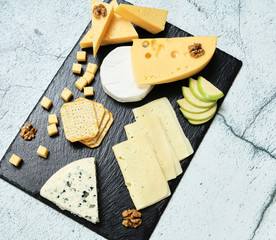 Pieces of cheese on a dark background. Cheeseboard. Sliced ​​apple and nuts on the board. Hard cheese, camembert, roquefort, emmental.