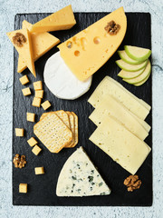 Pieces of cheese on a dark background. Cheeseboard. Sliced ​​apple and nuts on the board. Hard cheese, camembert, roquefort, emmental.