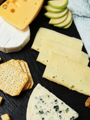 Pieces of cheese on a dark background. Cheeseboard. Sliced ​​apple and nuts on the board. Hard cheese, camembert, roquefort, emmental.