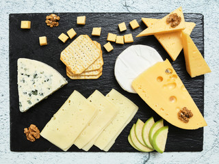 Pieces of cheese on a dark background. Cheeseboard. Sliced ​​apple and nuts on the board. Hard cheese, camembert, roquefort, emmental.