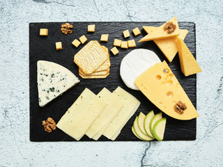 Pieces of cheese on a dark background. Cheeseboard. Sliced ​​apple and nuts on the board. Hard cheese, camembert, roquefort, emmental.