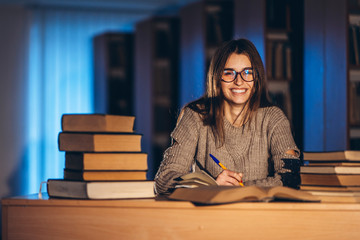 Young happy smiling student in glasses preparing for the exam. Girl in the evening sits at a table in the library with a pile of books, smiling and looking at the camera