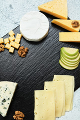 Pieces of cheese on a dark background. Cheeseboard. Sliced ​​apple and nuts on the board. Hard cheese, camembert, roquefort, emmental.