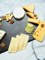 Pieces of cheese on a dark background. Cheeseboard. Sliced ​​apple and nuts on the board. Hard cheese, camembert, roquefort, emmental.