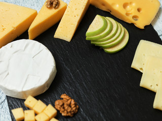 Pieces of cheese on a dark background. Cheeseboard. Sliced ​​apple and nuts on the board. Hard cheese, camembert, roquefort, emmental.