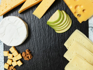 Pieces of cheese on a dark background. Cheeseboard. Sliced ​​apple and nuts on the board. Hard cheese, camembert, roquefort, emmental.