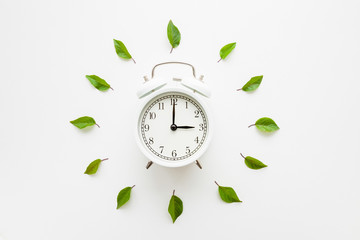 Green leaves around white alarm clock on light table background. Time change concept. Closeup. Flat lay. Top down view.
