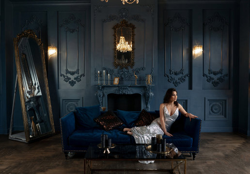 Magnificent brunette in silver shining dress impressive posing at sofa in chic royal interior