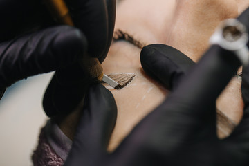 Hands in black gloves hold the manipulator with the microblading needle and draw her hair on the girl’s eyebrows close-up.