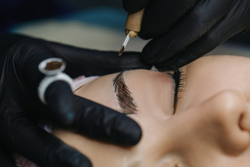 One hand in a black glove holds a manipulator with a microblading needle over the girl’s eyebrow, on the finger of the other hand is a pigment ring with a single cup closeup.