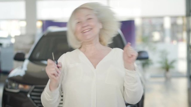 Portrait of excited mature Caucasian blond woman holding car keys, jumping and dancing in dealership. Cheerful senior businesswoman buying new vehicle in showroom. Joy, success, wealth, lifestyle.