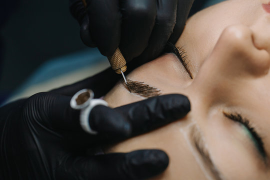 Hands in black gloves hold the manipulator with the microblading needle and draw her hair on the girl’s eyebrows close-up.