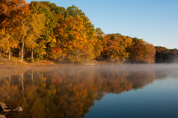 Fototapeta na wymiar The mist rolls into the bank of the lake and rises up to meet the brilliant autumn colors on a chilly morning.