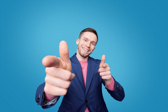 Portrait of cheerful successful young man in jacket making finger guns against blue background