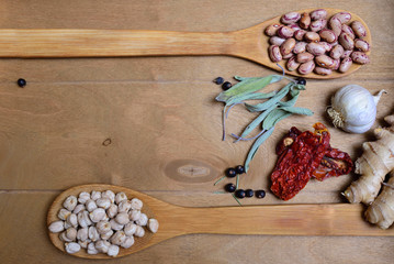Top view of a wooden table with various ingredients for cooking, such as a spoonful of beans, juniper berries, sun-dried tomatoes, ginger, garlic and sage leaves