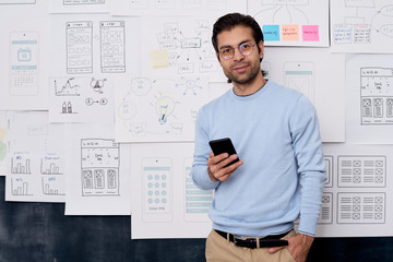 Horizontal medium portrait shot of young Caucasian man working in IT industry standing with smartphone looking at camera