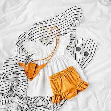 White Dress, Orange Shorts With Kids Handbag And Sunglasses. Set Of  Baby Clothes And Accessories For Summer Vacation On  Bed. Fashion Childs Outfit. Flat Lay, Top View