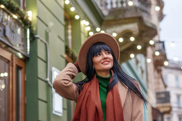 Outdoor close up portrait of young happy smiling brunette woman wearing classic beige hat, coat, scarf, brown suede gloves, walking in street of European city. Copy, empty space for text. 