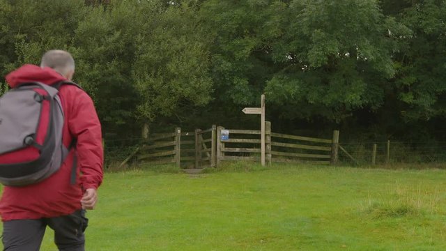 A daylight medium shot of a man wearing a red jacket and a backpack walking through a field towards the woods lined by an old rickety fence.