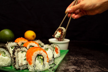 In a female hand, chopsticks hold sushi roll and soak in soy sauce. Sushi on a plate on a dark background with copy space. Tasting sushi rolls. Japanese food.