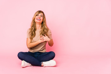 Young blonde caucasian woman sitting on a pink studio laughing keeping hands on heart, concept of happiness.