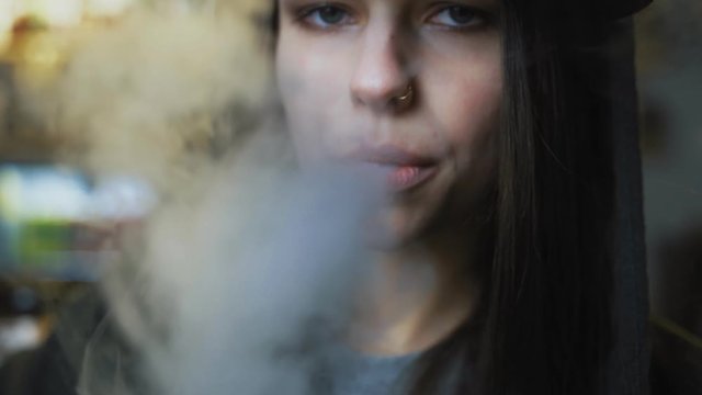 Young pretty woman in cap smoke an electronic cigarette at the vape shop. Hip-hop style. Closeup.