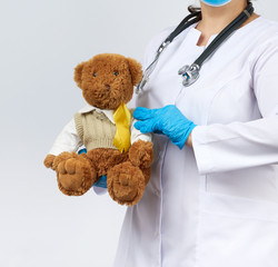 pediatrician in white coat, blue latex gloves holds a brown teddy bear with a yellow ribbon on a...