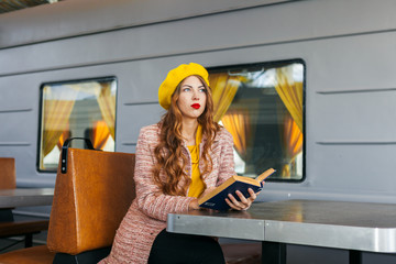 Portrait of awesome girl, wearing coat and yellow beret. Sitting on the train station read a book and looking straight. Autumn concept.