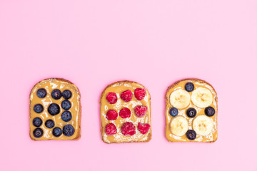 Top view of peanut butter sandwiches or toasts with banana, raspberry and blueberry on soft pink background with copy space. Healthy eating concept