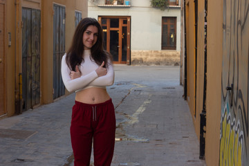 Portrait of positive caucasian young woman model keeps hands crossed and showing thumbs up gesture, in the city, white top and red pants. Place for your text in copy space.