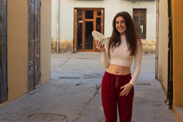 Portrait of pretty caucasian young woman model with dollar banknotes, in the city, white top and red pants, dark air. Place for your text in copy space.