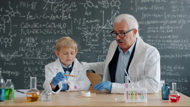 Little boy schoolchild is condusting chemical test at school while mature man teacher is helping explaining theory, board with formulas is in background.
