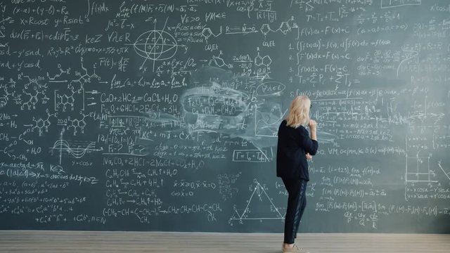 Time-lapse of female professor walking near chalkboard looking at formulas thinking looking for solution for scientific problem. People and knowledge concept.