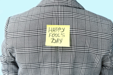 Sticky note with text HAPPY FOOL'S DAY on back of woman against color background. April Fools' Day...