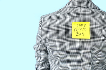 Sticky note with text HAPPY FOOL'S DAY on back of woman against color background. April Fools' Day...