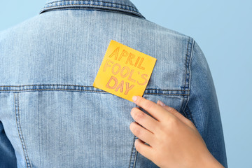 Woman sticking paper to her friend's back against color background, closeup. April Fools' Day prank