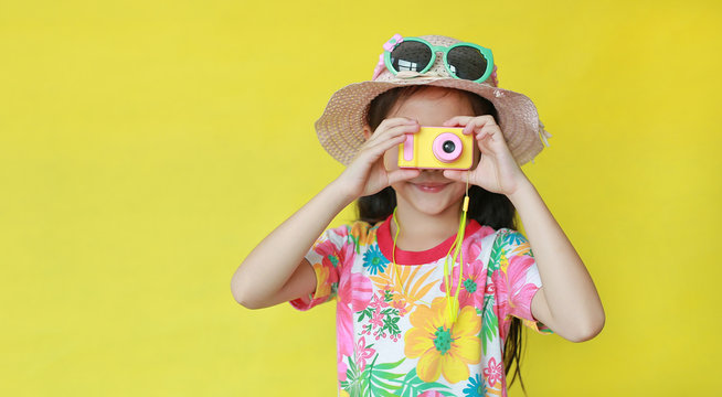 Little photographer taking photo by toy camera. Child tourists in floral pattern summer dress and hat with sunglasses isolated on yellow background