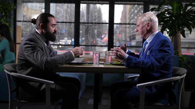 Respectable formally dressed businessmen commuticating at lunch in cozy city restaurant. Old man with gray hair and younger partner eating, discussing issuses of mutually beneficial cooperation