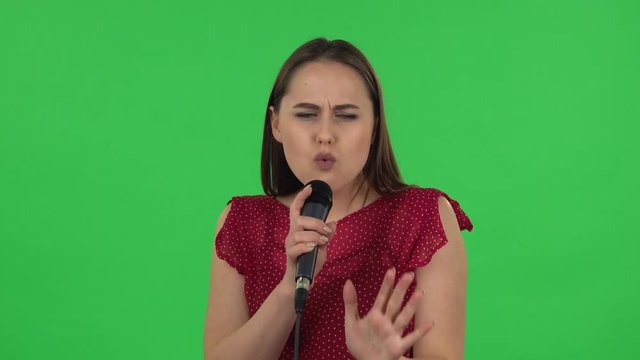 Portrait of tender girl in red dress is singing into a microphone and moving to the beat of music. Green screen
