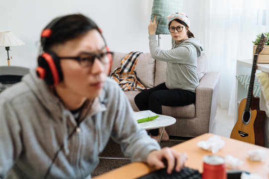 bokeh view of young asian lazy guy in headphones enjoys playing online computer game and girl on sofa angry with boyfriend holding pillow ready to throw. frustrated woman frowning staring at boy.