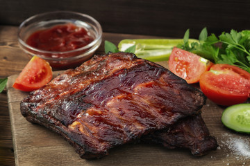 Barbecue pork ribs on a wooden cutting board with ketchup and vegetables