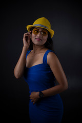 Fototapeta premium Fashion portrait of an young and attractive Indian Bengali brunette girl with blue western dress and yellow fashionable cap in front of black studio background. Indian fashion portrait and lifestyle.