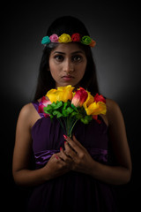 Fashion portrait of young and attractive Indian Bengali brunette girl in purple western dress and flower head band with vibrant flowers in hand in front of black studio background. Indian lifestyle.