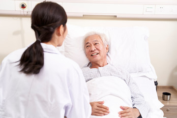 asian old man lying in bed talking to young female doctor in hospital ward
