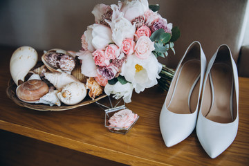 On a wooden table are white shoes, next to them is a wedding bouquet of flowers and greens, a glass box for engagement rings and a vase with shells
