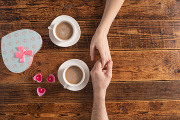 Valentine's Day celebration concept. A nice gift for your loved one. Hands of man and woman with coffee mugs on a wooden table background. Copy space. Flat lay. Close-up.