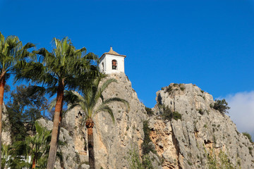 Fototapeta na wymiar white bell tower of castell de guadalest with palms on bright blue sky background, Spain