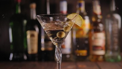 Close-up of a toothpick with olives falling into a Martini glass at the bar on the bar counter against the background of various bottles. Vermouth and gin cocktail with lime and olives at the bar. - Powered by Adobe